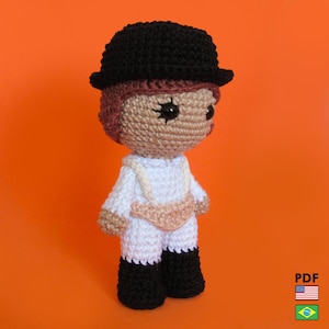 Op de afbeelding: Gehaakte pop in de stijl van een filmpersonage. De pop heeft een zwarte bolhoed, een witte jumpsuit en zwarte laarzen. De huid van de pop is bruin en het haar is roodbruin. De achtergrond is oranje.