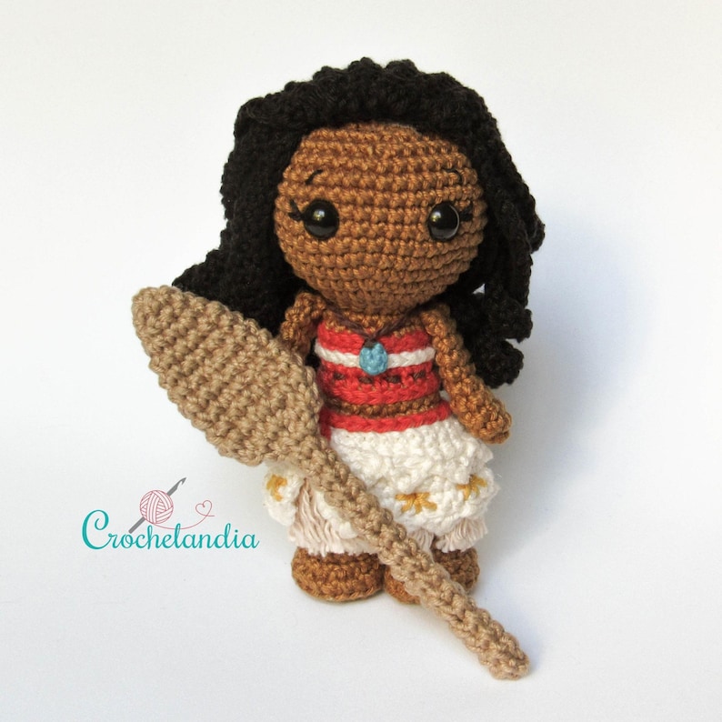 May include: Crocheted doll of a character with dark skin, black hair, and a red and white striped top. The doll is holding a tan crocheted paddle. The doll is wearing a white skirt with yellow accents. The doll is handmade.