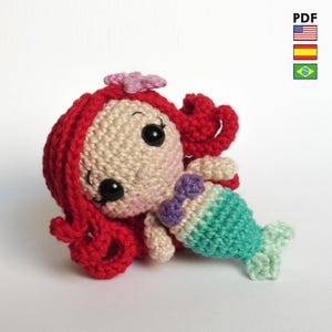 May include: A crochet amigurumi mermaid doll featuring bright red yarn hair with a small pink flower, a beige face with black safety eyes, a purple bikini top, and a teal green crocheted tail. The doll is presented as a PDF pattern file.