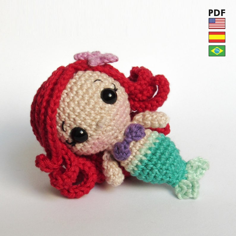 May include: A crochet amigurumi mermaid doll featuring bright red yarn hair with a small pink flower, a beige face with black safety eyes, a purple bikini top, and a teal green crocheted tail. The doll is presented as a PDF pattern file.
