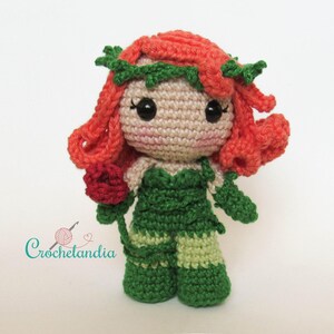 May include: A handmade crochet doll with red hair and a green outfit, holding a red rose. The doll has large black eyes and a smiling expression. The doll's hair is styled with green leaves. The text "Crochetlandia" is in the lower left corner.