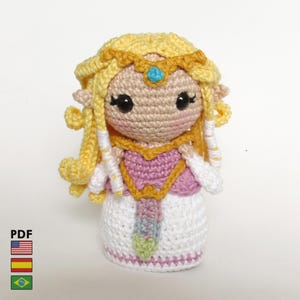Può includere: Bambola all'uncinetto fatta a mano raffigurante un personaggio con capelli biondi, una corona d'oro e un vestito rosa e bianco. La bambola ha grandi occhi neri e una piccola gemma blu sulla corona. La bambola è alta circa 15 cm.