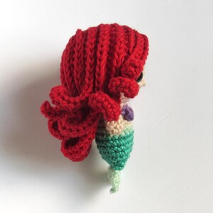 May include: A small crocheted mermaid doll featuring vibrant red yarn hair styled in thick, textured waves. The figure has a beige-coloured torso, a purple shell-shaped top, and a green crocheted tail that tapers into a light green fin.
