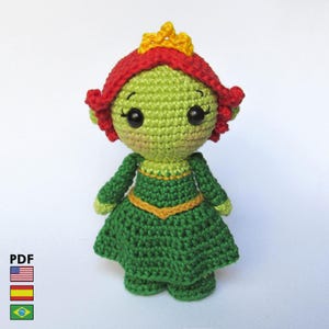 Puede incluir: Muñeca de ganchillo hecha a mano con cuerpo verde, pelo rojo y corona amarilla. La muñeca lleva un vestido verde con ribete amarillo. La muñeca tiene ojos negros y está sobre una superficie blanca. La imagen incluye el texto "PDF" y banderas.