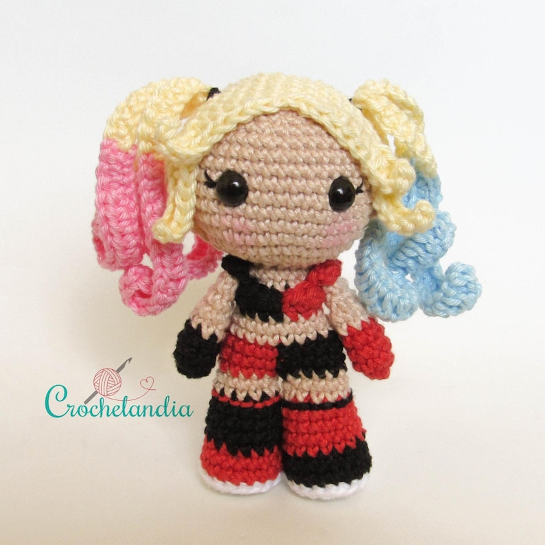 May include: A handmade crochet doll with blonde pigtails, one pink and one blue. The doll wears a black, red, and white checkered outfit with a red bow. The doll has large black eyes and a small pink nose. The text "Crochetlandia" is in the lower left corner.