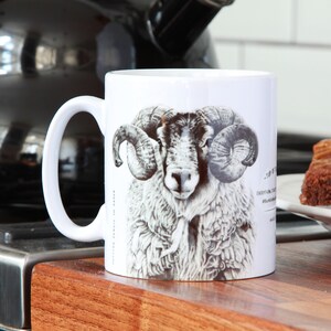 Peut inclure: Une tasse en céramique blanche avec une illustration en noir et blanc d'un mouton avec de grandes cornes. La tasse est posée sur une surface en bois.