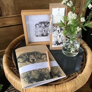 Pu&ograve; includere: Un taccuino marrone con un'illustrazione in bianco e nero di un ariete sulla copertina. Il taccuino &egrave; su un tavolo di vimini con un vaso di vetro di fiori bianchi e un segnalibro con un'illustrazione di ariete. Il segnalibro recita "Ewe Gotta Read This!" e "Bookmarker".