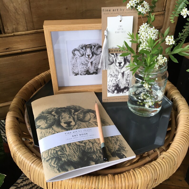 Pu&ograve; includere: Un taccuino marrone con un'illustrazione in bianco e nero di un ariete sulla copertina. Il taccuino &egrave; su un tavolo di vimini con un vaso di vetro di fiori bianchi e un segnalibro con un'illustrazione di ariete. Il segnalibro recita "Ewe Gotta Read This!" e "Bookmarker".