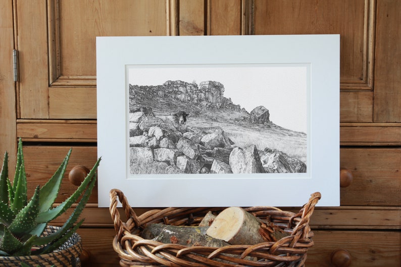 Pu&ograve; includere: Un disegno in bianco e nero di un paesaggio roccioso con una mucca in piedi in primo piano. Il disegno &egrave; incorniciato in una cornice bianca ed &egrave; esposto su una superficie di legno.