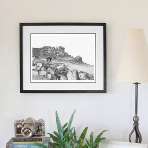 Pu&ograve; includere: Fotografia in bianco e nero di un paesaggio montuoso roccioso incorniciata da una cornice nera. La fotografia &egrave; appesa a un muro bianco con una lampada e una macchina fotografica su uno scaffale sottostante.