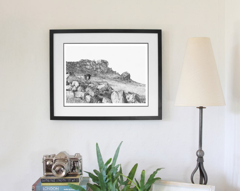 Pu&ograve; includere: Fotografia in bianco e nero di un paesaggio montuoso roccioso incorniciata da una cornice nera. La fotografia &egrave; appesa a un muro bianco con una lampada e una macchina fotografica su uno scaffale sottostante.