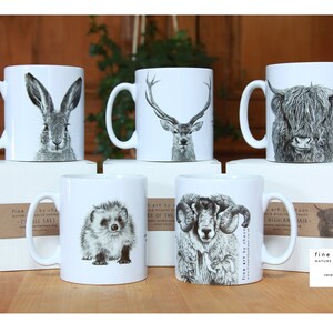 Pu&ograve; includere: Sei tazze in ceramica bianca con illustrazioni in bianco e nero di vari animali, tra cui una lepre, un cervo, una mucca delle Highlands, un riccio, un ariete e un'altra mucca delle Highlands. Le tazze sono disposte in due file da tre, con la fila inferiore che poggia su scatole con etichette di carta marrone che recitano "fine art by shaun nature & wildlife portraits ceramic earthenware mug made local".