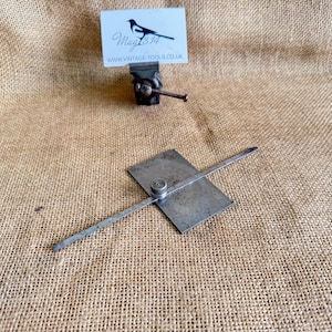 May include: An antique metal tool with a rectangular base and a pivoting arm, likely used for measuring or marking. A small vise and a business card with a bird logo are also visible. The items rest on a textured, tan fabric.