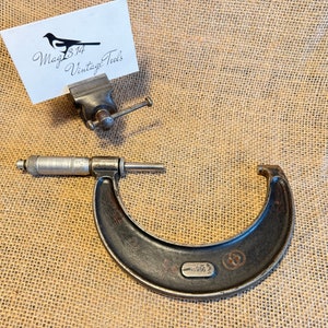 May include: A vintage black and silver micrometer with a measurement of 1.056. The micrometer is on a hessian background.