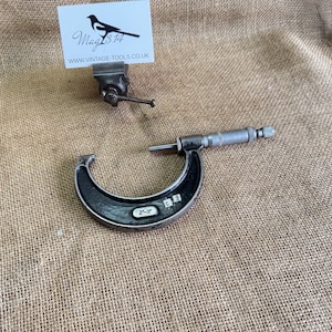 May include: A vintage micrometer with a black and silver finish, measuring 2-3 inches. The tool is set against a textured, tan background. A small vise and a business card with a bird logo and website address are also visible.