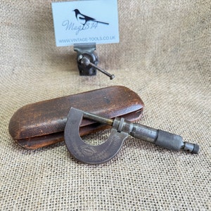 May include: A vintage, silver-coloured, outside micrometer with a wooden case. The micrometer has a knurled adjustment knob and a graduated scale. The case is brown and has a hinged lid.