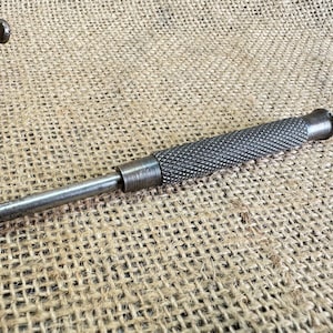 May include: A metal punch tool with a textured handle and a pointed tip. The tool is designed for making holes in metal.