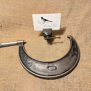 May include: A vintage, silver-coloured, outside micrometer with a black handle. The micrometer is on a hessian background. The micrometer is labelled "Mitutoyo".