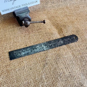 Puede incluir: Un tornillo de banco de metal antiguo y una regla con un agujero en un extremo, ambos con signos de desgaste. El tornillo de banco es de color gris oscuro con un mango plateado. La regla es de color gris oscuro y tiene marcas de medición. Los artículos descansan sobre una superficie de arpillera.