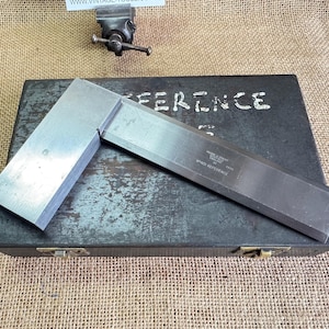 May include: A vintage steel machinist's square on a dark metal box. The square is a right angle tool for precise measurements. The box is marked with the word "REFERENCE". A small vise is visible in the background. The square is a tool for engineering.