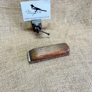 Könnte beinhalten: Ein Holz-Schaber mit einer Metallklinge. Der Schaber ist braun und hat eine rechteckige Form. Die Klinge ist silberfarben und mit einer Metallschraube am Schaber befestigt.