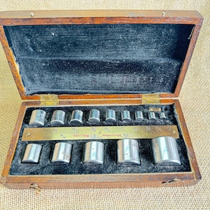 May include: A wooden box containing a set of 14 metal weights. The weights are labelled in fractions of an inch, from 1/16 to 1 1/4. The box is labelled "J.H.J. HOPFMANN, 1 ROLLERS, 3/16"