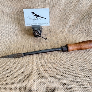 May include: A vintage wood-handled metal chisel with a pointed tip. The chisel is lying on a hessian surface. A small vice is in the background with a business card that reads "Mag 814 www.vintage-tools.co.uk".