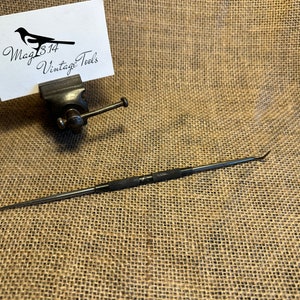 Puede incluir: Una herramienta de metal negro con una punta puntiaguda y un mango texturizado. La herramienta mide aproximadamente 15 cm de largo y está sobre una superficie de arpillera marrón. La herramienta está etiquetada como "Mag 814 Vintage Tools".