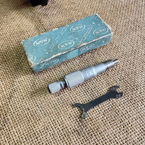 May include: A vintage M&W micrometer with its original box and a small wrench. The micrometer is silver, and the box is teal with the M&W logo. The wrench is black. The items are on a burlap surface.