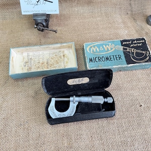 May include: A silver micrometer in a black case, alongside a blue and white box marked "M&W MICROMETER", rests on a burlap surface. An open, light blue box with text is also present. The micrometer is a precision measuring tool.