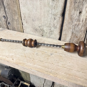Op de afbeelding: Een vintage handboorgereedschap met een lange, gedraaide metalen schacht, een donkerbruin houten handvat aan het uiteinde en een kleinere, bijpassende verschuifbare houten greep. De metalen schacht is afgewerkt met een kleine moer en messingkleurige accenten.