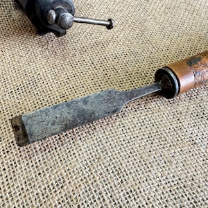 Könnte beinhalten: Ein antiker Holzmeißel mit einer Klinge aus Metall und einem Holzgriff. Der Meißel liegt auf einer Jutestoff-Oberfläche.