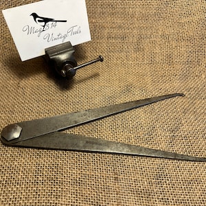 May include: A vintage metal compass with a pointed tip and a hexagonal head. The compass is lying on a hessian surface.
