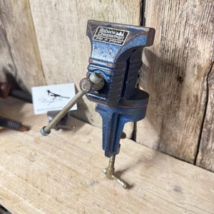 May include: A blue metal bench vise with a silver handle and a screw mechanism for mounting. The top of the vise features a black and white label with the text Rabone Chesterman Made in England. The tool has visible signs of wear and rust.