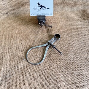 May include: An antique metal outside caliper and a small vice are displayed on a burlap surface. The caliper has a curved design with an adjustable screw. A business card with a magpie logo and website address is in the background.