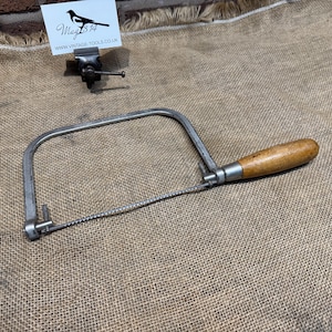 May include: A vintage coping saw with a wooden handle and a metal frame. The saw blade is visible, and the handle is a light brown colour. The saw is resting on a burlap surface. A small sign with a bird logo and text is in the background.