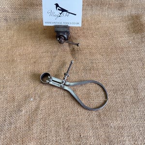 May include: A vintage metal caliper, a tool for measuring, with a curved design. The silver-toned tool is displayed on a textured, tan fabric. A small vice and a business card with a bird logo are also visible.