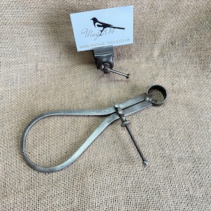 May include: A vintage metal caliper tool with a curved design and adjustable screw. A small card with a bird logo and the text "Mag 314" and "www.VINTAGE-TOOLS.co.uk" is held in a small vice. The background is a textured, beige fabric.