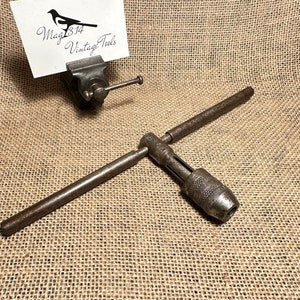 Puede incluir: Un portabrocas de taladro vintage de metal con un mango en forma de T. El portabrocas está oxidado y tiene un agarre moleteado. El mango está hecho de metal y tiene un agarre de madera.