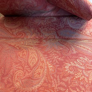 May include: Close-up of a paisley fabric in shades of red, peach, and cream. The fabric features a detailed, repeating pattern of curved, teardrop-shaped motifs and floral elements. The fabric is rolled up, revealing the pattern.