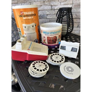 May include: A selection of vintage View-Master items, including a red and white talking View-Master, a white viewer, and several reels. The packaging includes the text "TALKING VIEW-MASTER GIFT PAK II" and "VIEW-MASTER Gift Pak Cartoon Favourites."