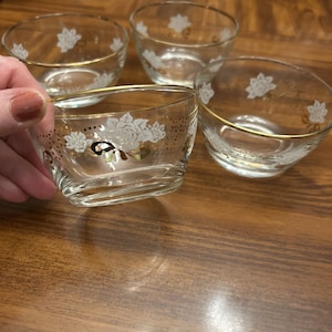 May include: Four clear glass bowls with gold rims and white floral designs. One bowl is being held by a hand. The bowls are on a wooden surface. The floral design includes roses and other decorative elements.