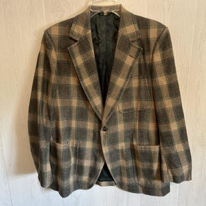 May include: A blazer featuring a tan and dark gray plaid pattern. The garment includes a single-button front closure, notched lapels, and two large patch pockets on the lower front, with a black interior lining visible inside the open jacket.