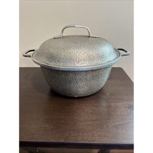 Op de afbeelding: Een zilverkleurige, getextureerde metalen kookpot met een bijpassende deksel. De pot heeft twee zijhandvatten en een enkele handgreep op het deksel. De pot staat op een donkerbruin houten oppervlak.