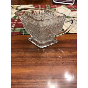May include: A clear pressed glass creamer pitcher featuring a textured diamond pattern on the exterior, a square base, a curved handle, and a pouring spout. The glass surface displays intricate geometric detailing and a raised relief design.