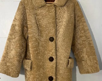 May include: A tan sheepskin coat with a brown button closure and two pockets. The coat is hanging on a wooden hanger.
