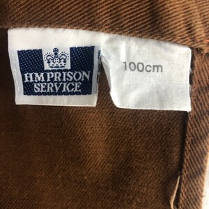 Pode incluir: Close-up de um tecido castanho com uma etiqueta que diz "HM PRISON SERVICE" e uma etiqueta com "100cm" impressos. O tecido tem um aspeto texturizado.