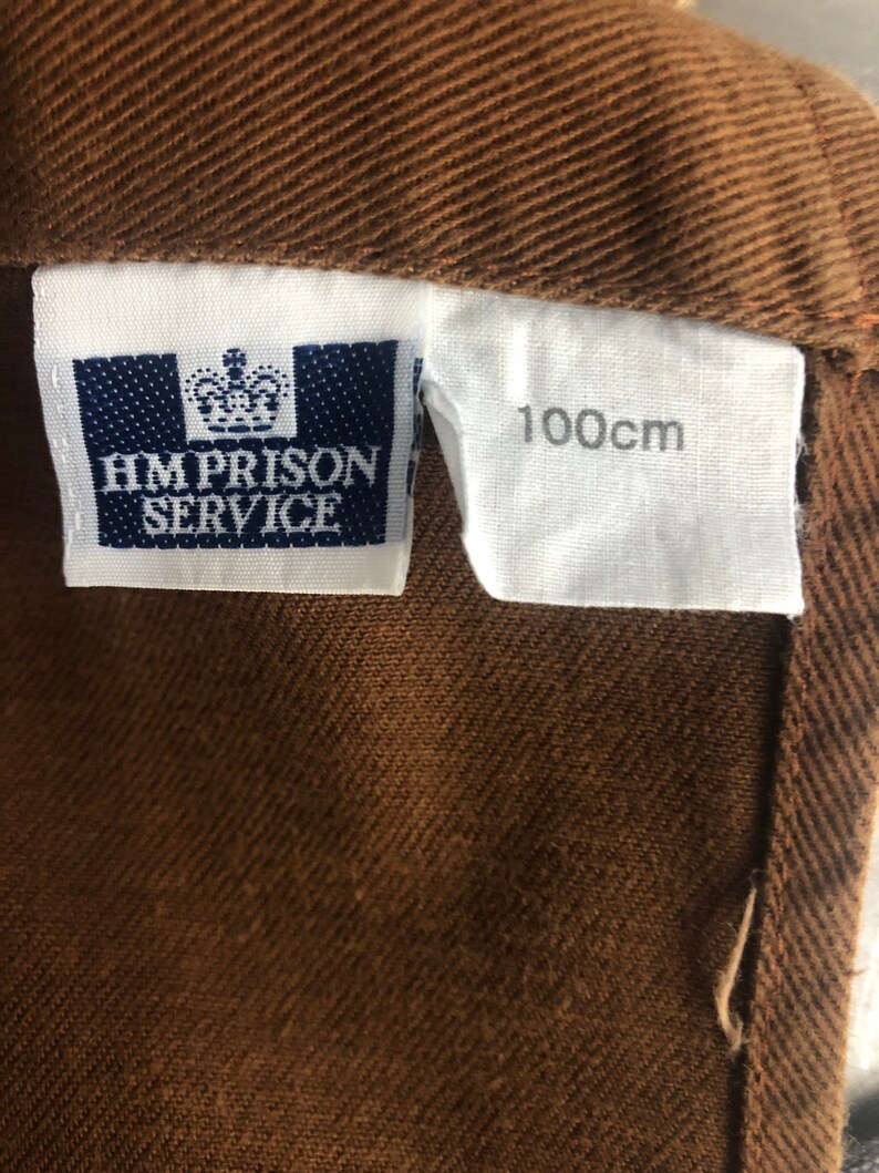Pode incluir: Close-up de um tecido castanho com uma etiqueta que diz "HM PRISON SERVICE" e uma etiqueta com "100cm" impressos. O tecido tem um aspeto texturizado.