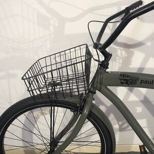 May include: A grey bicycle with a black wire basket, black handlebars, and a black tyre. The bicycle has a front mudguard and a logo on the frame. The bicycle is parked in front of a white wall.