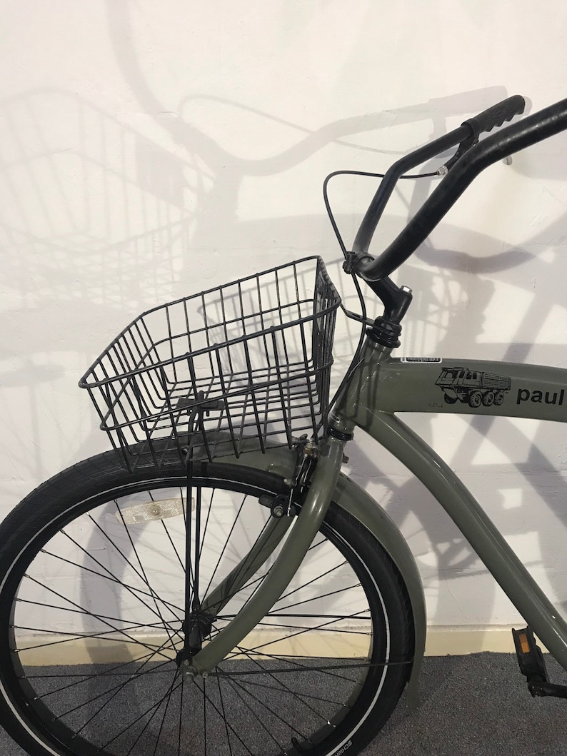 May include: A grey bicycle with a black wire basket, black handlebars, and a black tyre. The bicycle has a front mudguard and a logo on the frame. The bicycle is parked in front of a white wall.